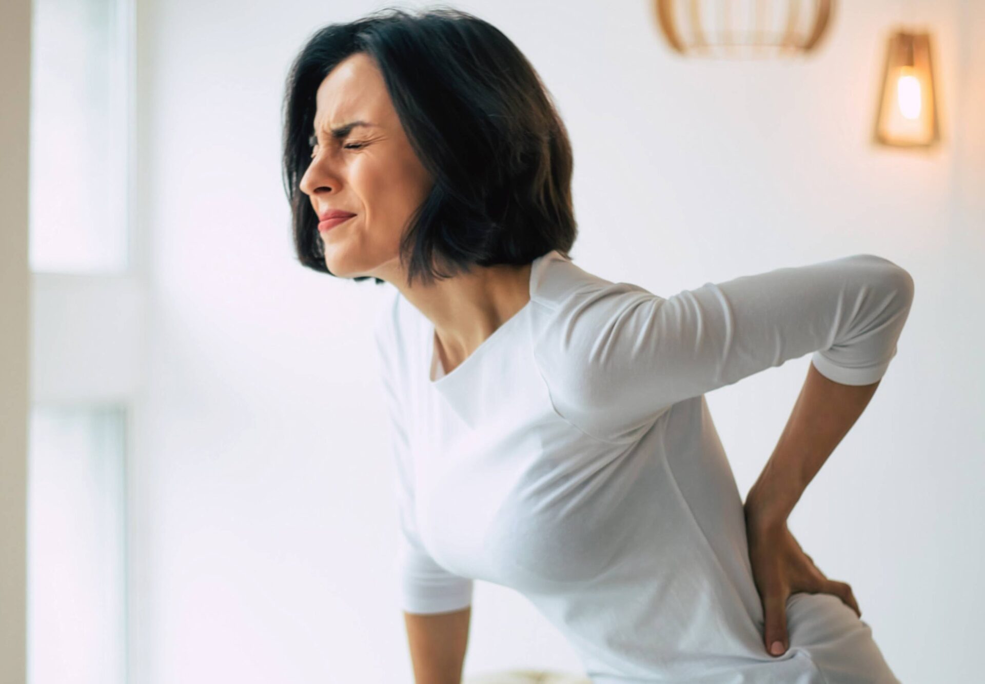 A woman at home, grimacing as she supports her lower back, depicting the struggle with back pain