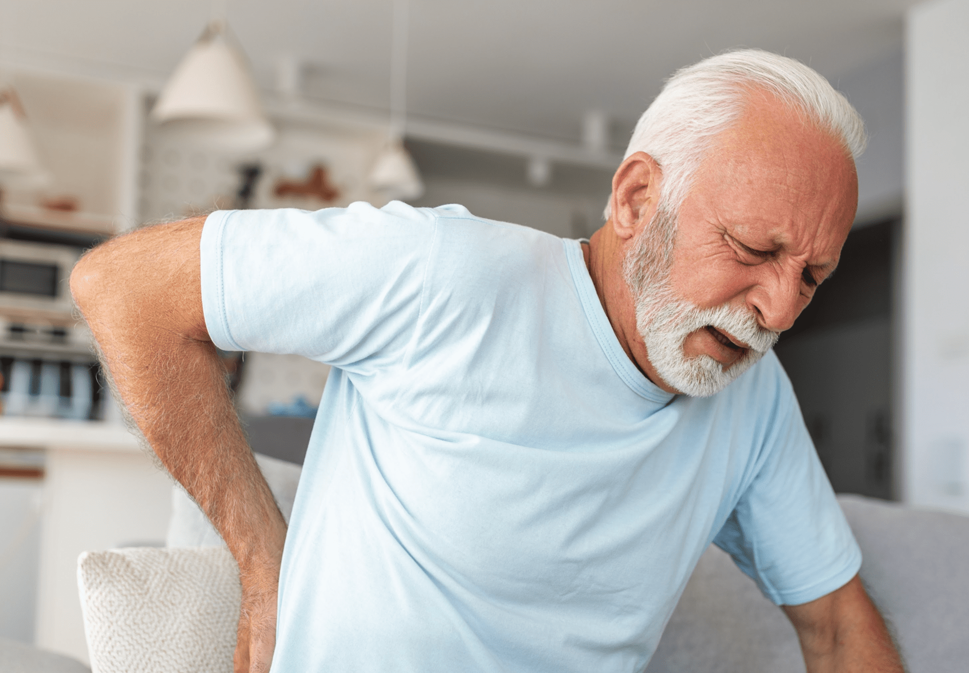 A man in his living room, grimacing as he experiences back pain, with his hand resting on his lower back