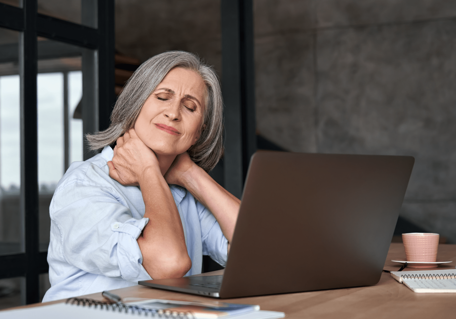 A woman seated at a desk, visibly uncomfortable, holding her neck in pain while working on her laptop